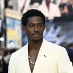Damson Idris in cream suit with gold chain, walking outdoors at a public event” Damson Idris in cream suit with gold chain, walking outdoors at a public event”