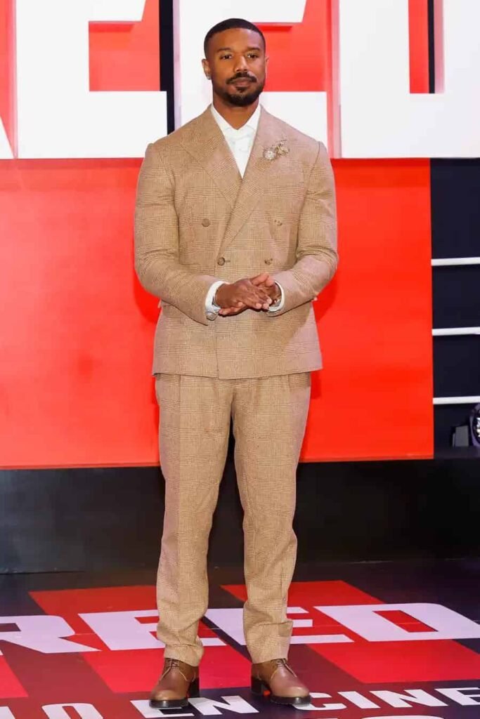 Michael B. Jordan camel double-breasted suit Michael B. Jordan wearing a camel double-breasted suit with brown dress shoes, posing on the red carpet in front of a bold backdrop