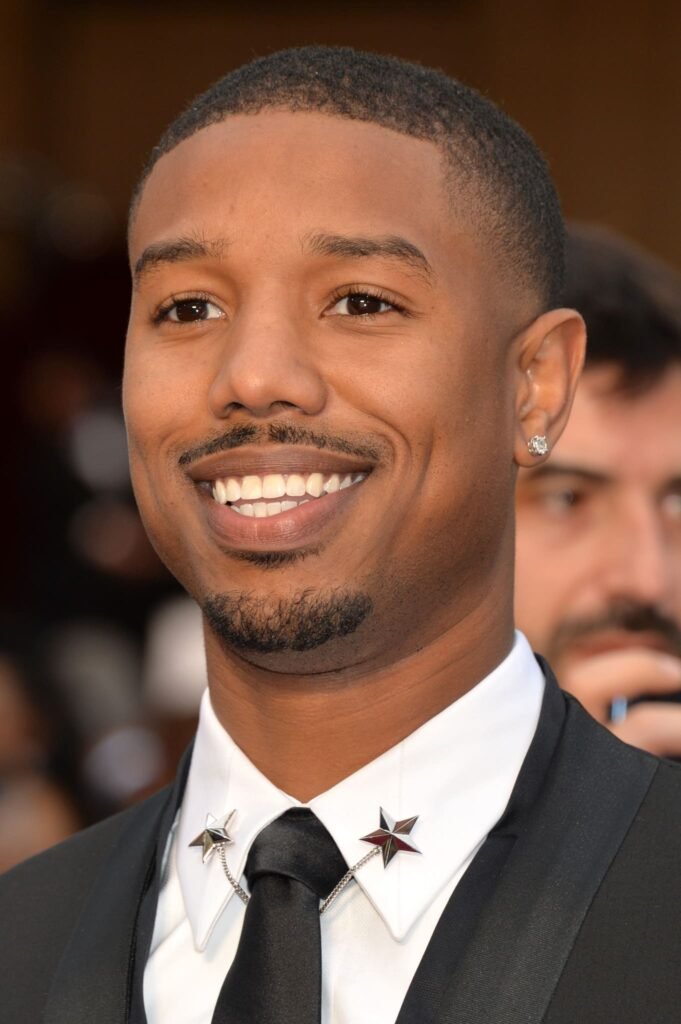 Michael B. Jordan in sleek tuxedo with star collar clips Michael B. Jordan smiling in a sharp black tuxedo featuring unique star-shaped collar pins at a formal red carpet event