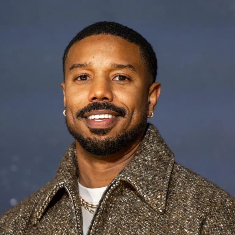 Michael B. Jordan in GQ-style wool jacket with gold chain Michael B. Jordan smiling in a textured brown wool jacket layered with a chain and white shirt on a blue backdrop