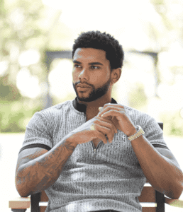 full haircut – outdoor look Stylish Black man with a full head and trimmed beard sitting outside in a grey shirt