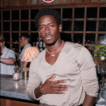 Damson Idris in a beige V-neck long sleeve shirt and dark trousers, styled with a gold watch and layered necklaces at a bar Damson Idris in a beige V-neck long sleeve shirt and dark trousers, styled with a gold watch and layered necklaces at a bar