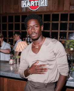 Damson Idris in a beige V-neck long sleeve shirt and dark trousers, styled with a gold watch and layered necklaces at a bar Damson Idris in a beige V-neck long sleeve shirt and dark trousers, styled with a gold watch and layered necklaces at a bar