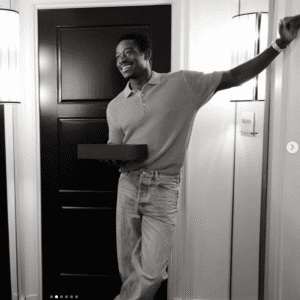 Damson Idris smiling in a doorway wearing a polo shirt and jeans, holding a gift box Damson Idris smiling in a doorway wearing a polo shirt and jeans, holding a gift box
