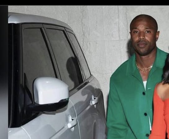 Clean Shaven Look Michael B. Jordan stepping out of a white SUV in a green jacket, sporting a clean-shaven face and a gold chain