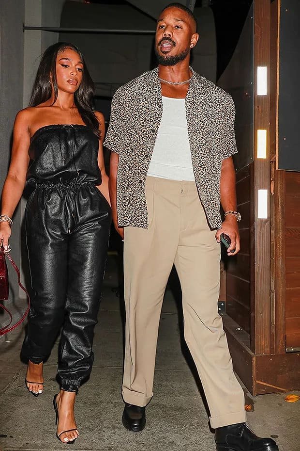 Michael B. Jordan date night outfit Michael B. Jordan wearing a patterned short-sleeve shirt over a white tee with beige trousers and black boots, walking beside a woman in a black leather strapless jumpsuit