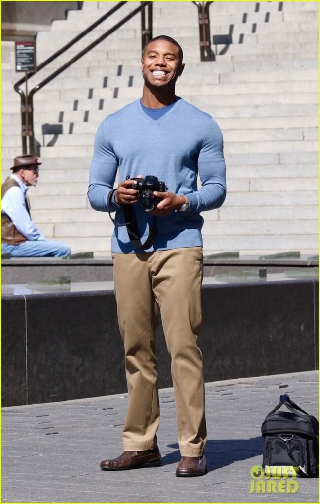 Michael B. Jordan casual fall outfit Michael B. Jordan wearing a fitted blue crewneck sweater with khaki trousers and brown loafers, holding a camera outdoors with steps and people in the background