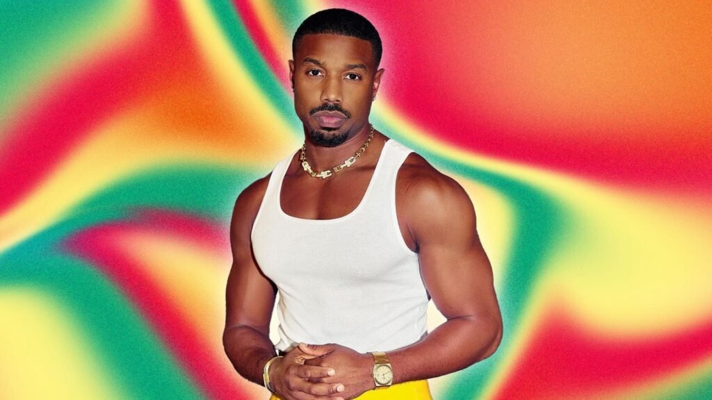 Michael B. Jordan tank top editorial Michael B. Jordan wearing a white tank top and gold watch, posing in front of a vibrant, colorful swirl background